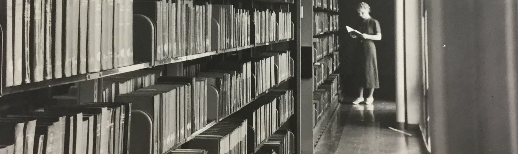 Lady looking through a book, with bookshelves in the foreground. Circa 1950.